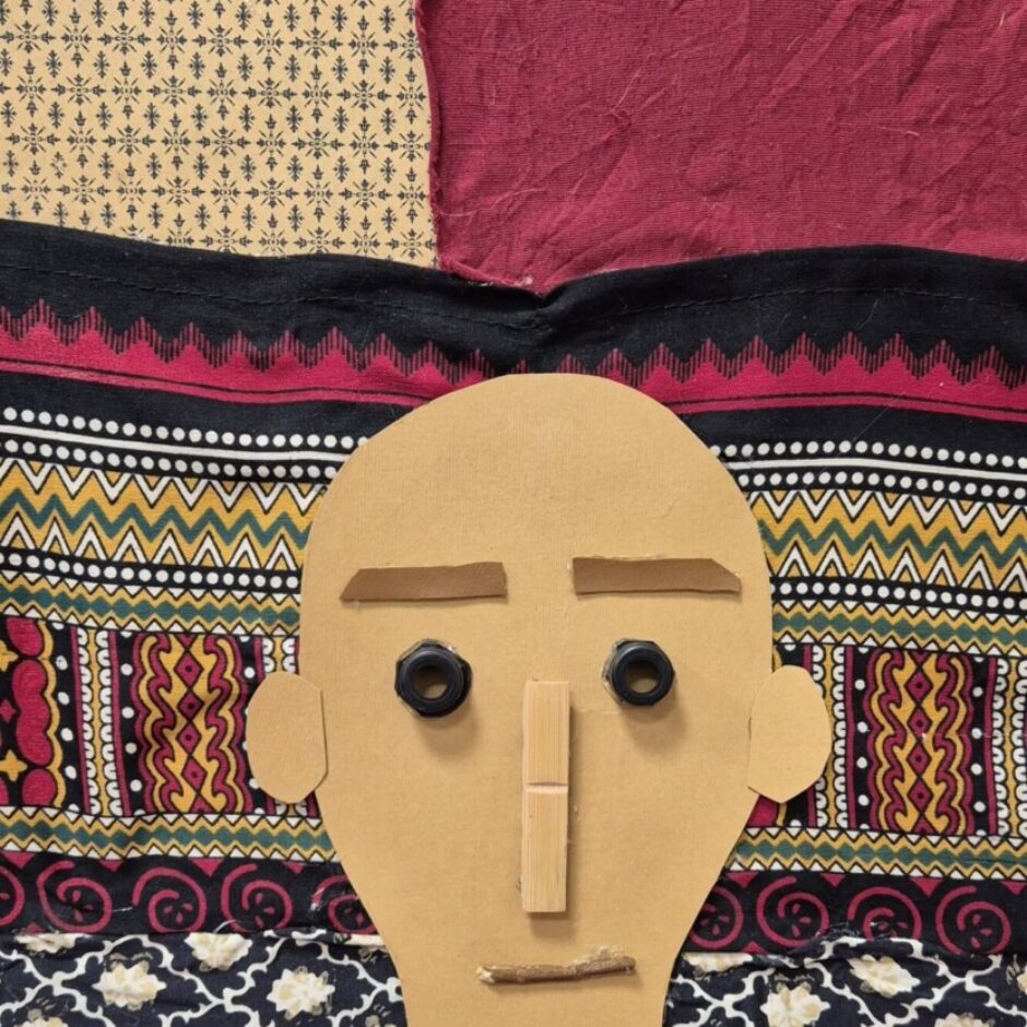 Kolāža ar cilvēka seju, veidota no auduma, pogām, papīru un dabas elementiem.