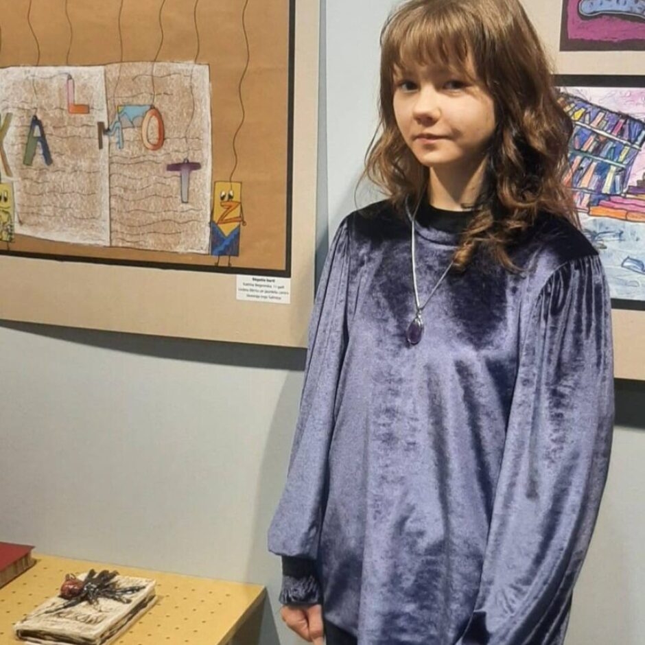 Jauna meitene ar gariem, brūniem matiem un poniju, ģērbusies tumši violetā samta blūzē un melnās biksēs, stāv mākslas izstādē.