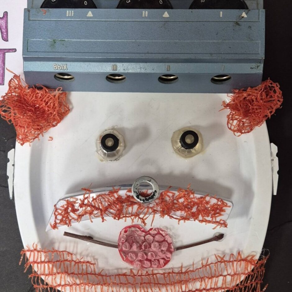 Kolāža ar cilvēka seju, veidota no auduma, pogām, papīru un dabas elementiem.