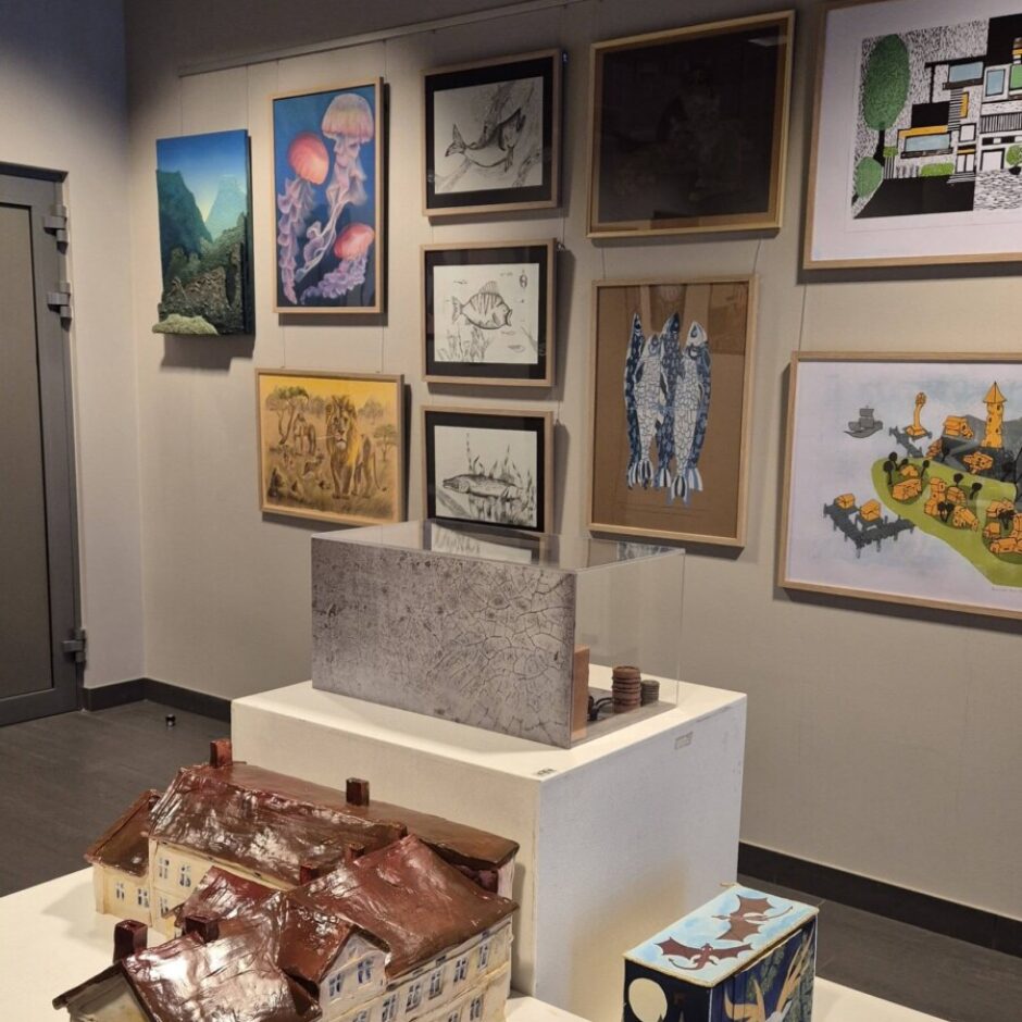 Mākslas izstāde ar daudzveidīgiem mākslas darbiem. Priekšplānā uz baltiem pjedestāliem izstādīti divi detalizēti ēku modeļi ar sarkanbrūniem jumtiem, plakans pelēks abstrakts panelis un neliela krāsaina kārba ar mēness, staltbrieža un sikspārņu attēlu. Pie sienas pakārti dažādi gleznojumi un zīmējumi, tostarp medūzu, zivju, Āfrikas savannas ainavas, modernas arhitektūras un stilizētu ciematu attēli, kā arī trīsdimensiju kalnu ainava.