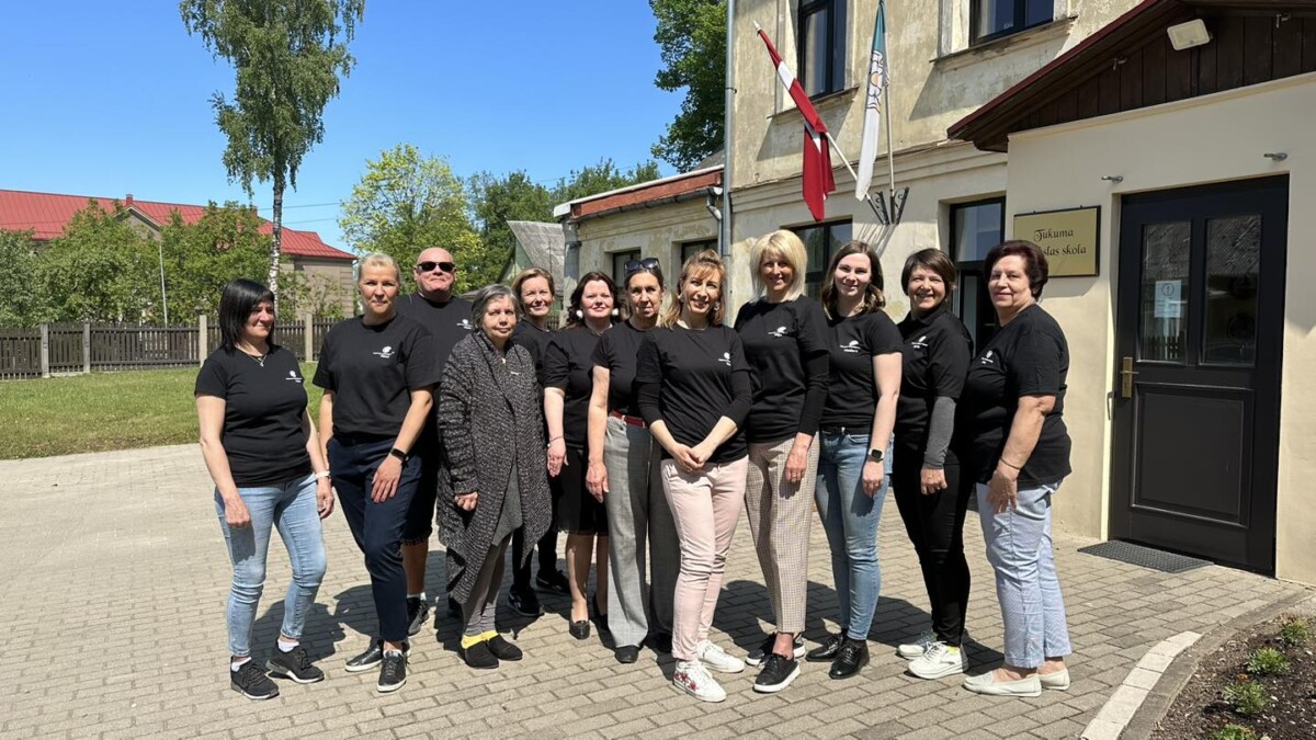 Tukuma Mākslas skolas kolektivs - divpadsmit cilvēki, pārsvarā sievietes, melnos T-kreklos ar logotipu, smaidoši pozē grupveida fotogrāfijai saulainā dienā.