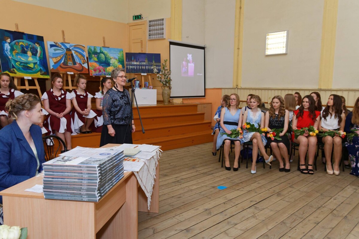 Sieviete ar brillēm un rakstainu blūzi runā mikrofonā, fonā stāv gleznas uz molbertiem un sēž divas studentu grupas: vienas bordo kleitās uz pakāpieniem, otras svinīgās kleitās ar ziediem rokās krēslos. Priekšplānā pie galda sēž sieviete, blakus liels grāmatu krāvums.