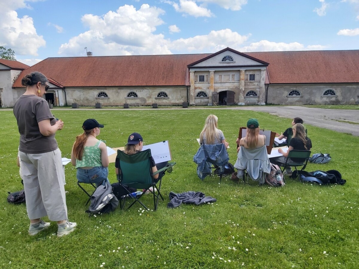 Grupa zīmē ārā uz zāles laukuma. Pieaugušais stāv un vēro, kamēr vairāki jaunieši sēž uz krēsliem un zīmē uz molbertiem vai blokiem.