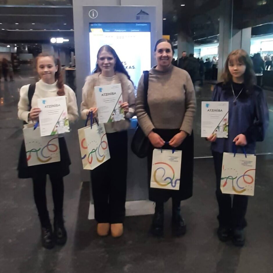 Trīs jaunas meitenes un viena sieviete smaidot pozē, katra turot baltu atzinības rakstu ar uzrakstu ATZINĪBA un baltu dāvanu maisiņu ar krāsainu rakstu, stāvot iekštelpās ar citiem cilvēkiem fonā.