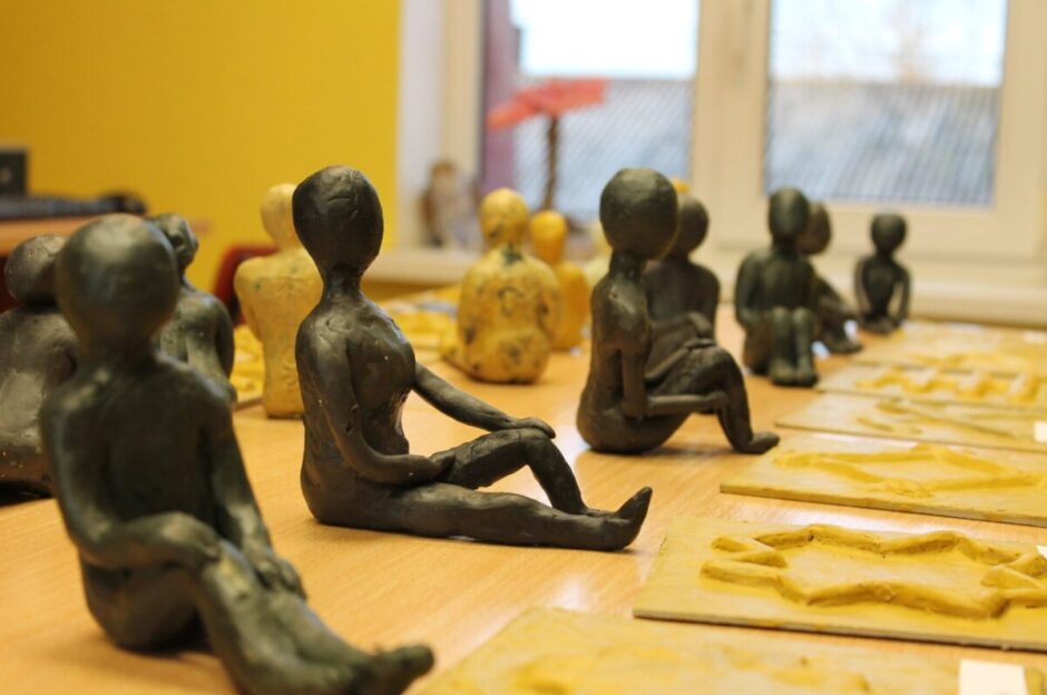 Uz koka galda izvietotas daudzas tumšas un dažas gaišas, cilvēkveidīgas māla figūriņas, kas sēž vai guļ.