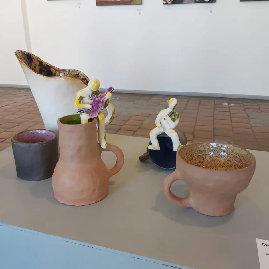 Uz pelēka pjedestāla izvietota keramikas kompozīcija ar piecām dažādu formu vāzēm un krūzēm, uz kurām novietotas divas stilizētas dzeltenīgi baltas figūriņas, kas spēlē instrumentus.