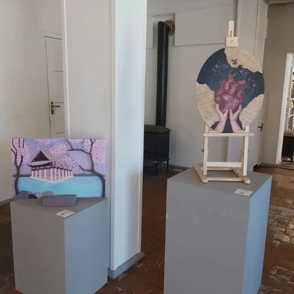 Divi mākslas darbi izstādīti iekštelpu galerijā. Kreisajā pusē uz pelēka pjedestāla ir glezna ar japāņu templi virs ūdens, ko ieskauj ķiršu ziedu koki. Labajā pusē uz koka molberta, kas novietots uz cita pelēka pjedestāla, ir apaļš mākslas darbs, kurā redzamas rokas, kas tur sirdi.